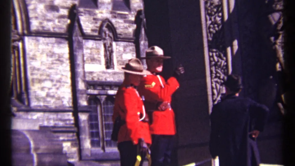 1967 Ottawa home-movie frame showing two RCMP officers beside a stone entrance as one gestures toward nearby visitors.