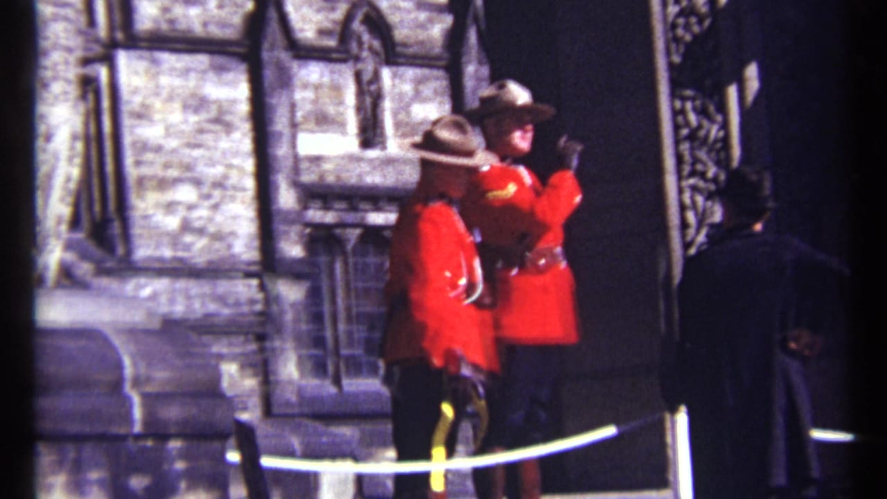 Two RCMP officers in red uniforms stand by a stone facade while one gestures with an arm raised.