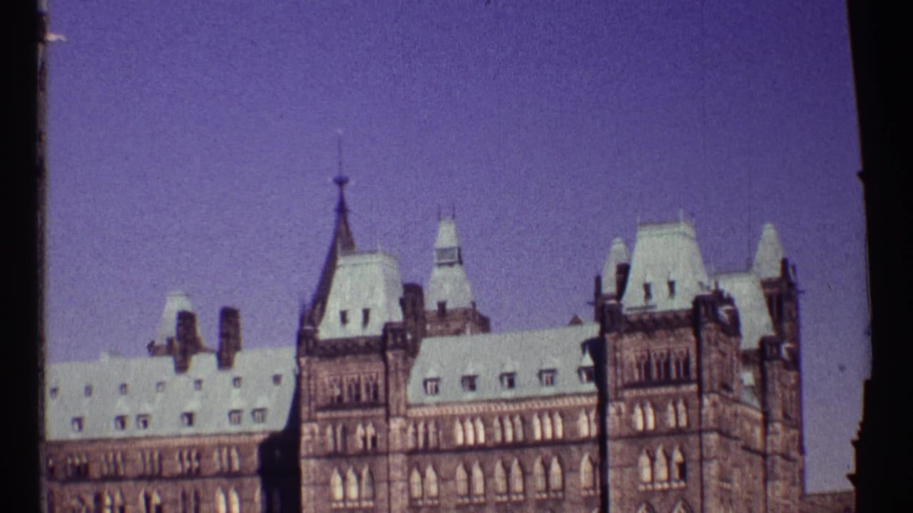 1967 Ottawa home-movie frame showing the upper Parliament Buildings with Gothic Revival towers and pale rooflines against a purple-tinted sky.
