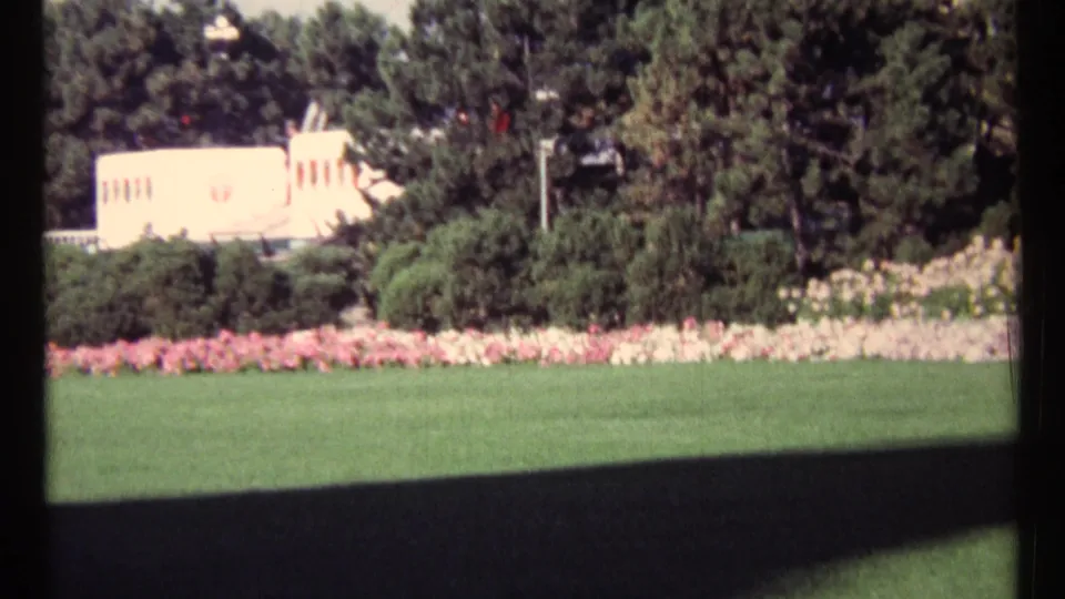 1967 Ottawa park view with a broad green lawn, pink and white flower beds, dense trees, and a pale structure partly visible behind plantings.