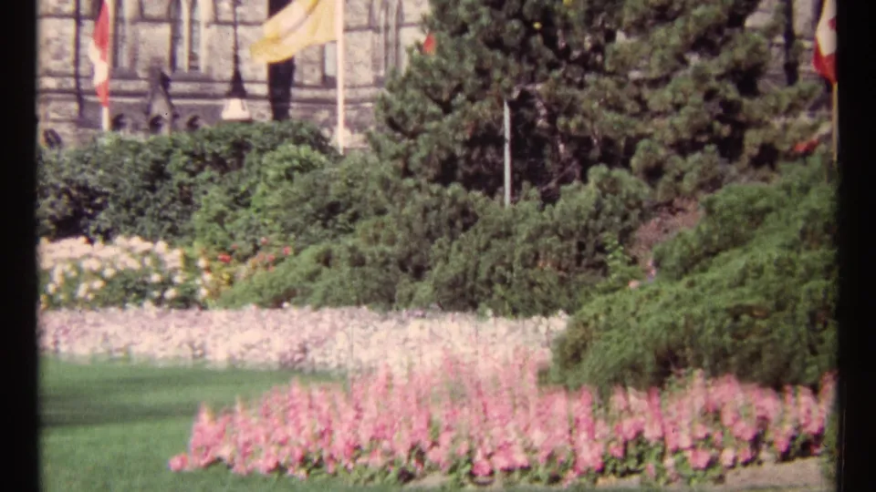 1967 Ottawa garden view with colorful flowerbeds, trimmed greenery, flagpoles, and a partial stone facade in the background.