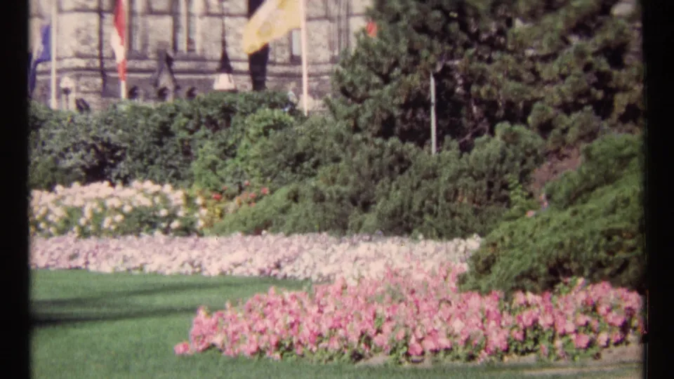1967 Ottawa frame of layered flowerbeds, hedges, conifers, and flagpoles in front of a partly visible stone building.