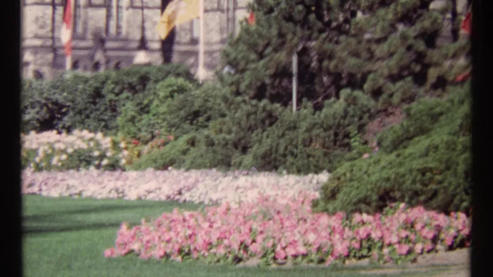 1967 Ottawa garden frame with bright flowerbeds, manicured shrubs, conifer trees, flagpoles, and a stone facade in soft 8mm color.