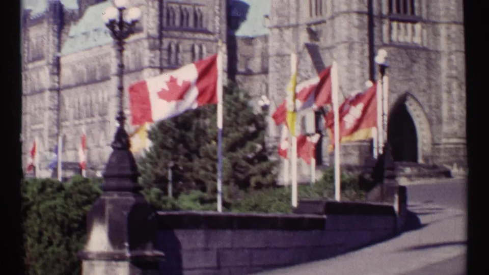 1967 Ottawa 8mm frame showing Canadian flags and other flagpoles in front of the Parliament Buildings, with a stone balustrade, shrubs, and an ornamental lamppost.