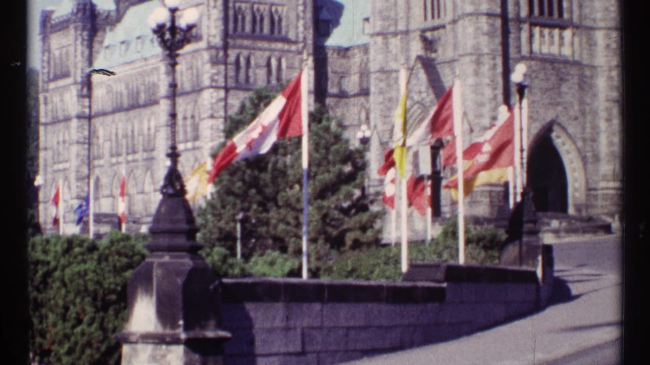 1967 Ottawa view of flags blowing near an ornamental lamppost, with the Parliament Buildings stone facade and arched entrance behind low greenery.