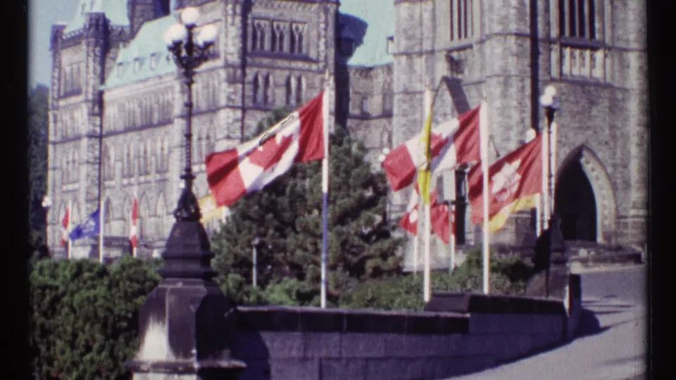 1967 Ottawa 8mm still with a large Canadian flag crossing the foreground, other flags beyond it, and the Parliament Buildings rising behind shrubs and stonework.