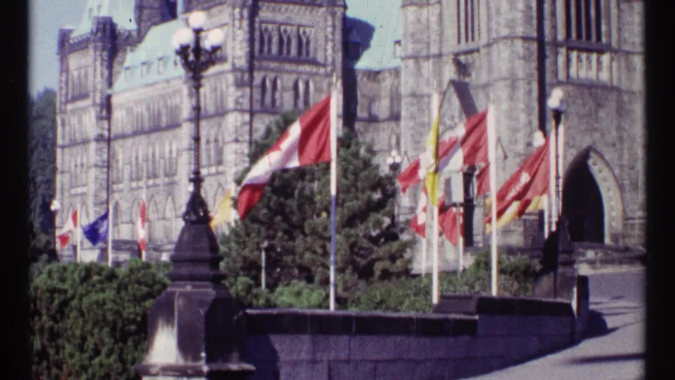 1967 Ottawa frame showing several flags in wind before the Parliament Buildings, with a dark lamppost, stone balustrade, shrubs, and arched doorway visible.