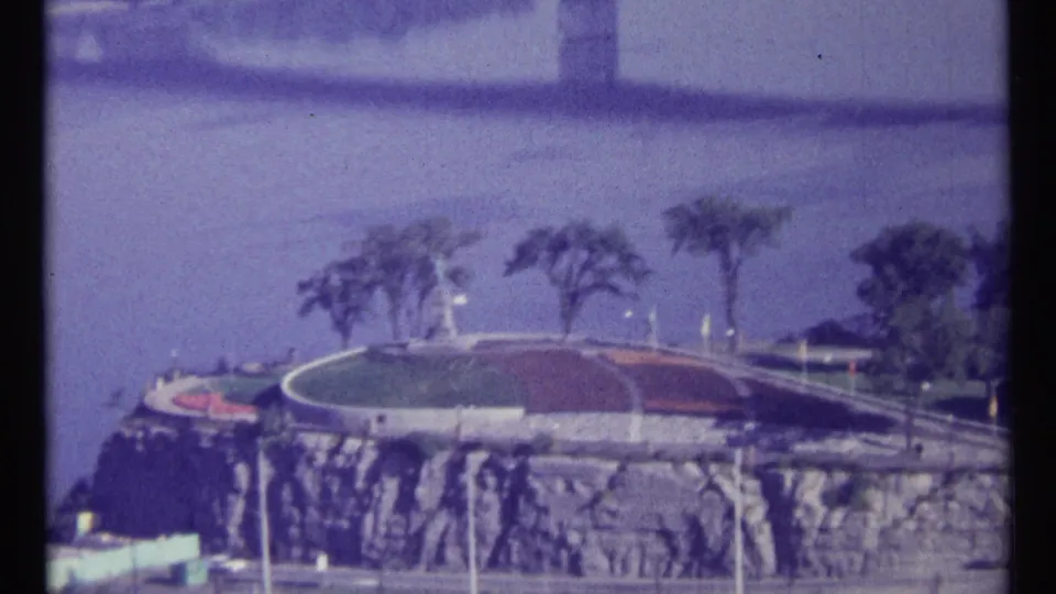 1967 Ottawa 8mm frame showing a riverfront overlook, rocky cliffside, landscaped circular park, road, trees, and bridge approach.