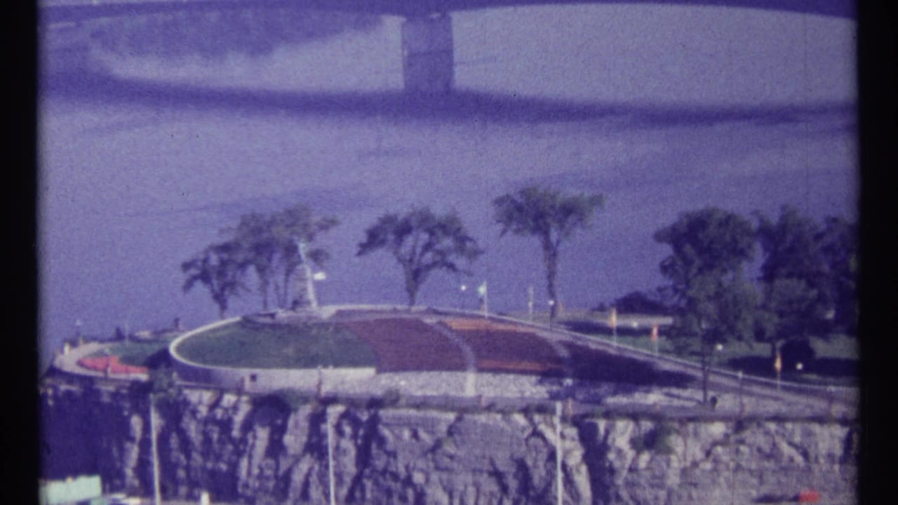 1967 Ottawa view across a riverfront cliff with a circular landscaped park, trees, road markings, and the bridge approach in soft 8mm color.