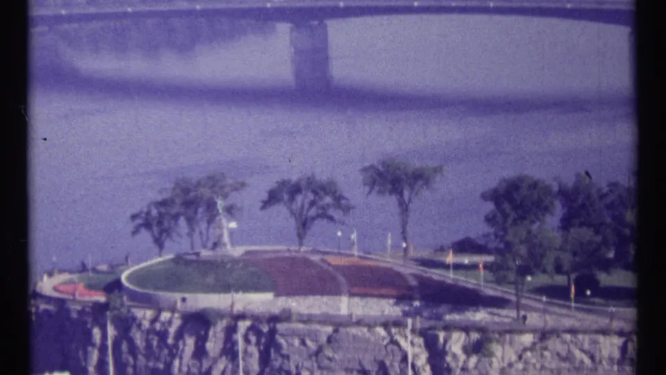Ottawa riverfront frame from 1967 showing a rocky embankment, landscaped grounds, road, trees, and muted water beyond in grainy 8mm film.