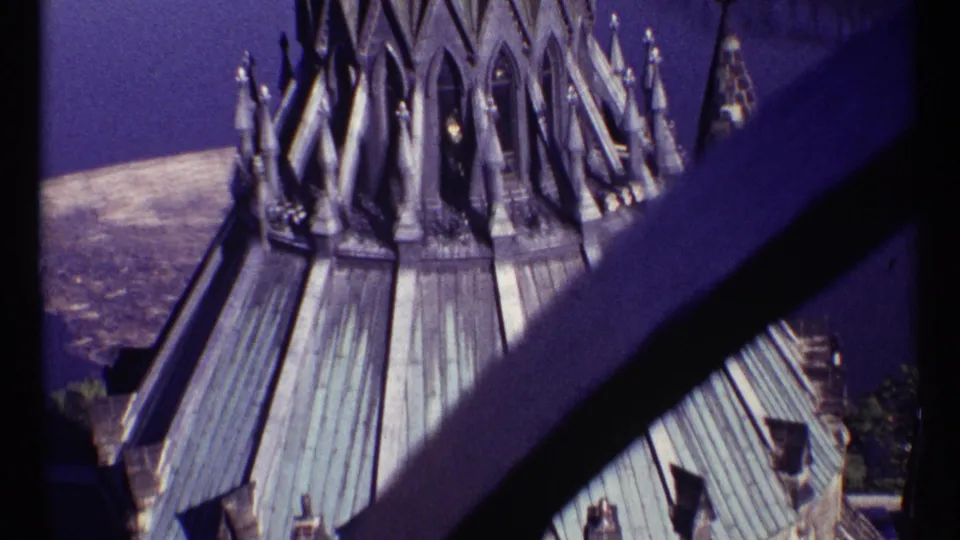 1967 Ottawa 8mm frame showing Parliament Hill Gothic roof details, Canadian flags, river water, and a dark bridge or railing crossing the foreground.