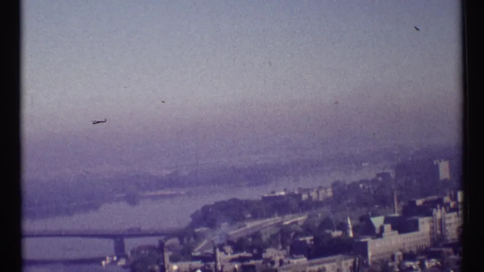 1967 Ottawa riverfront panorama with low-rise buildings, a shoreline road or path, trees, and a broad hazy river beyond.