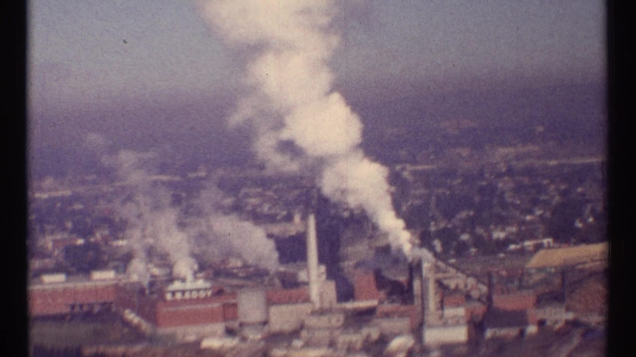 Industrial plant with multiple smokestacks releasing white smoke above a hazy cityscape.