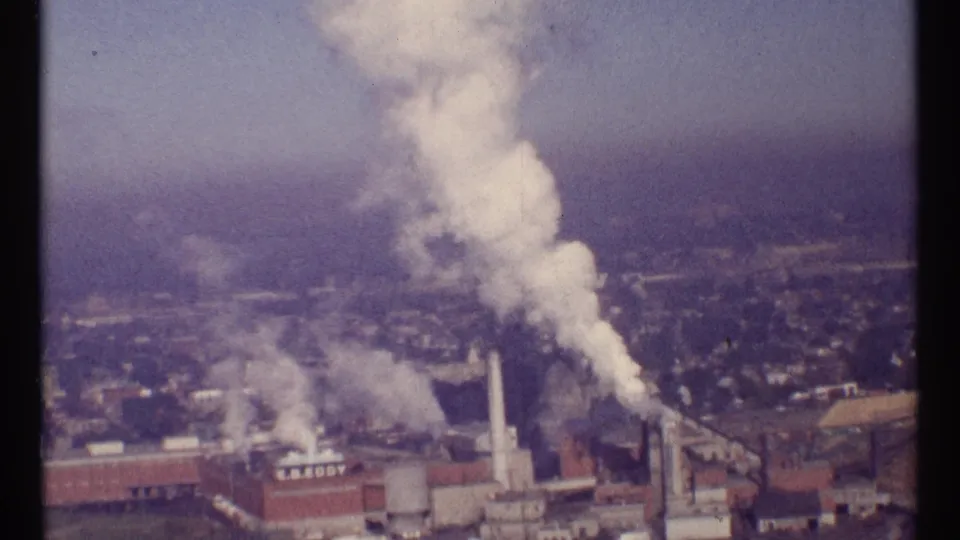 1967 Ottawa frame showing a tall smokestack and white smoke above an industrial site, with surrounding city blocks in haze.