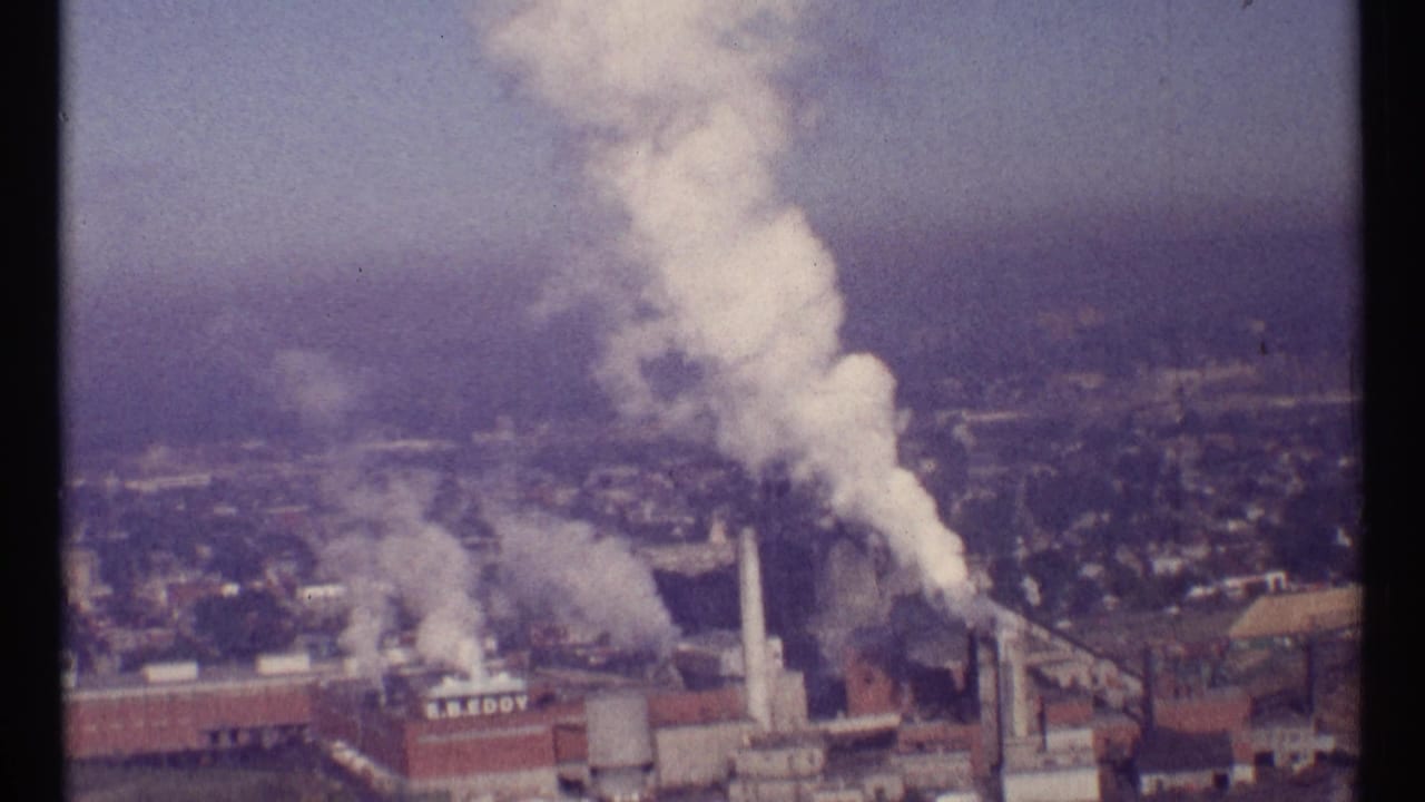 Smokestacks emit white smoke over industrial buildings in a hazy city view.
