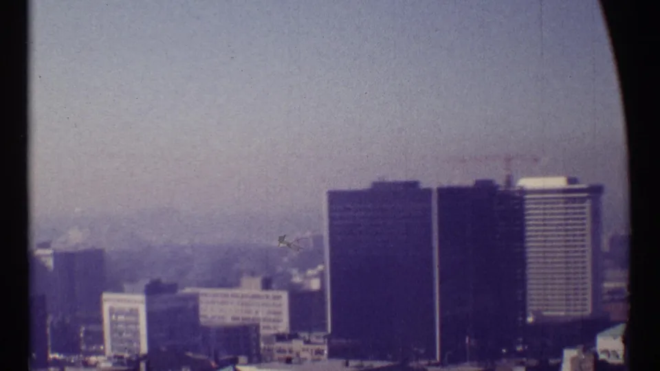 1967 Ottawa home-movie frame with dark high-rise blocks, a pale tower, and a rooftop crane under a washed pale sky.