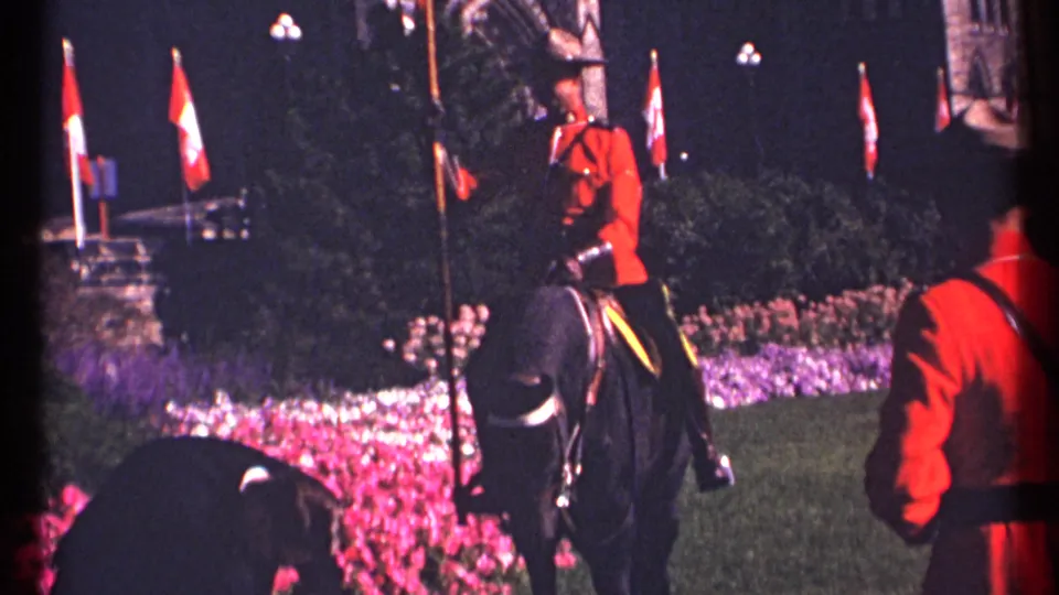 1967 Ottawa 8mm frame showing an RCMP mounted guard in red serge riding a black horse past lit flags and flowerbeds.