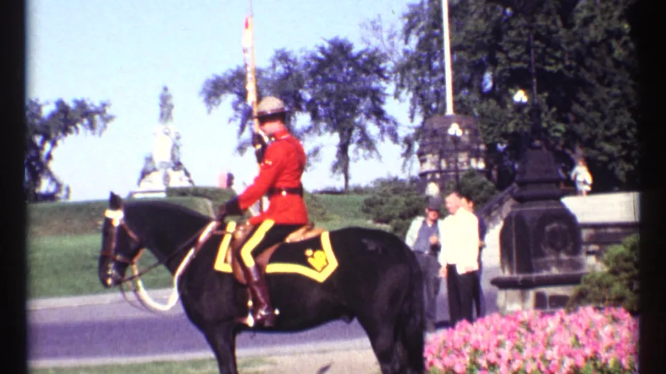 Daylight 1967 Ottawa frame showing an RCMP mounted guard on a black horse near flowerbeds, onlookers, and a distant monument.
