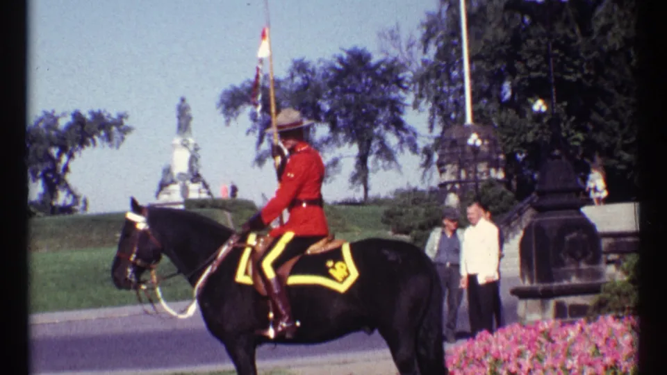 1967 Ottawa daylight frame of a red-serge RCMP rider on a black horse moving past flowerbeds, onlookers, and park monuments.