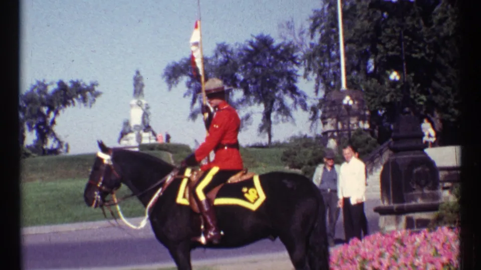 1967 Ottawa home-movie frame showing a mounted RCMP officer in red serge beside a road, with bystanders and a stone building nearby.