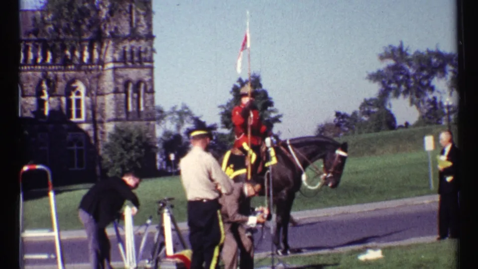 Ottawa 1967 home-movie frame showing the mounted RCMP officer, nearby personnel, camera equipment, and a suited onlooker beside the road.
