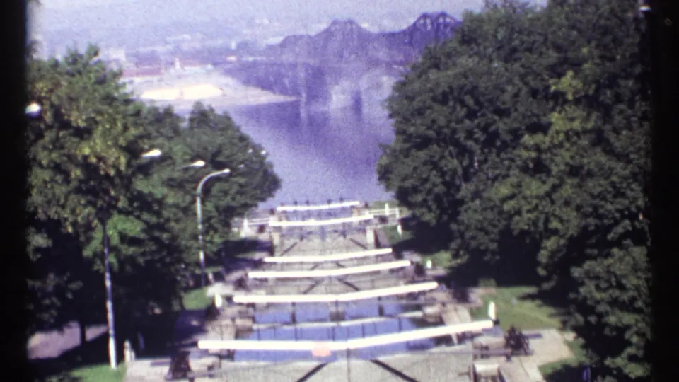 1967 Ottawa 8mm frame looking down a landscaped waterfront stairway toward water, trees, and distant hazy hills or mountains.