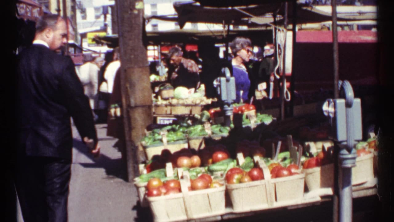Man in a dark suit walks past a sidewalk vegetable stand with baskets of tomatoes and greens.