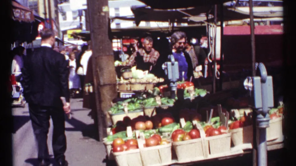 Rear view of a man in a dark suit walking past an Ottawa vegetable stand with baskets of tomatoes and greens in 1967 home-movie footage.