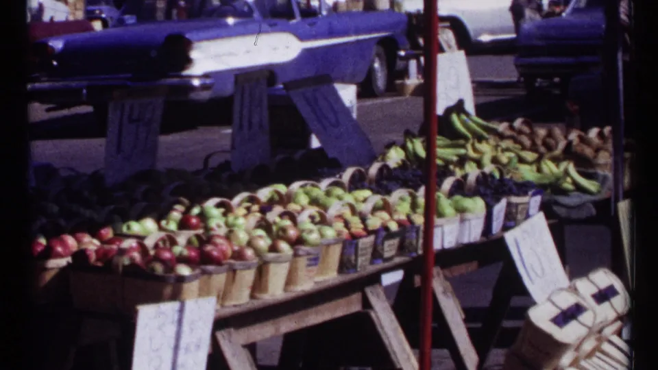 Ottawa market stall with apples, bananas, handwritten signs, wooden supports, and mid-century cars visible on the street behind.