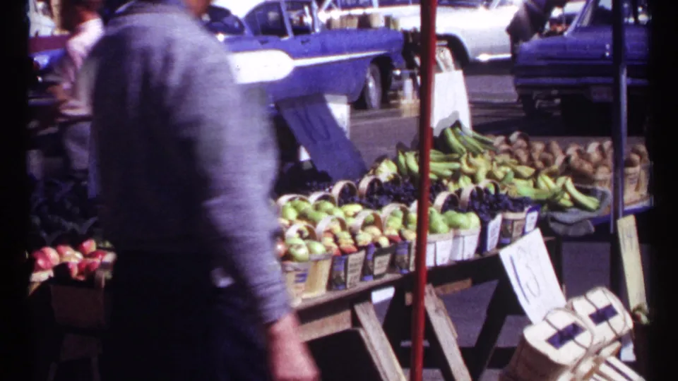 A passerby moves through the foreground of the 1967 Ottawa market scene, partly obscuring fruit baskets, bananas, signs, and parked cars.