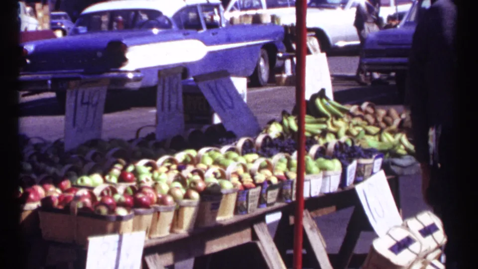 Curbside Ottawa produce display with apples, bananas, price signs, a red pole, wooden stands, and a prominent blue mid-century car behind.