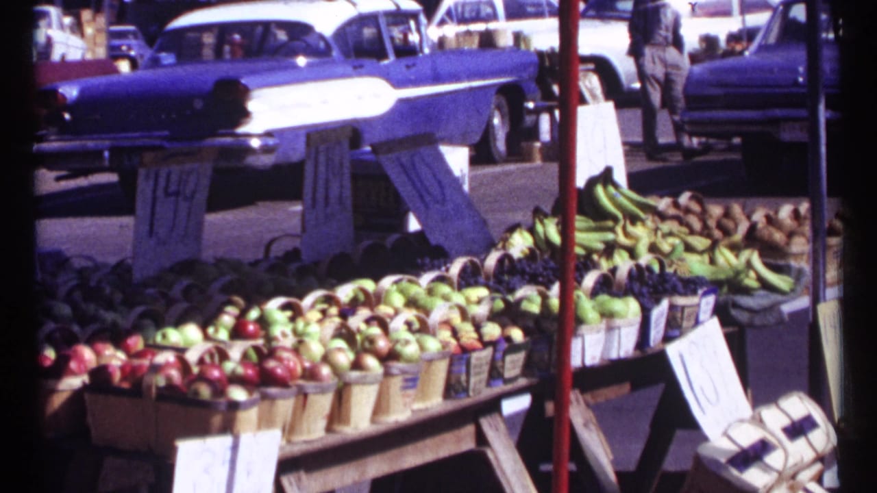 Outdoor produce stand with baskets of apples and bananas beside parked mid-century cars.