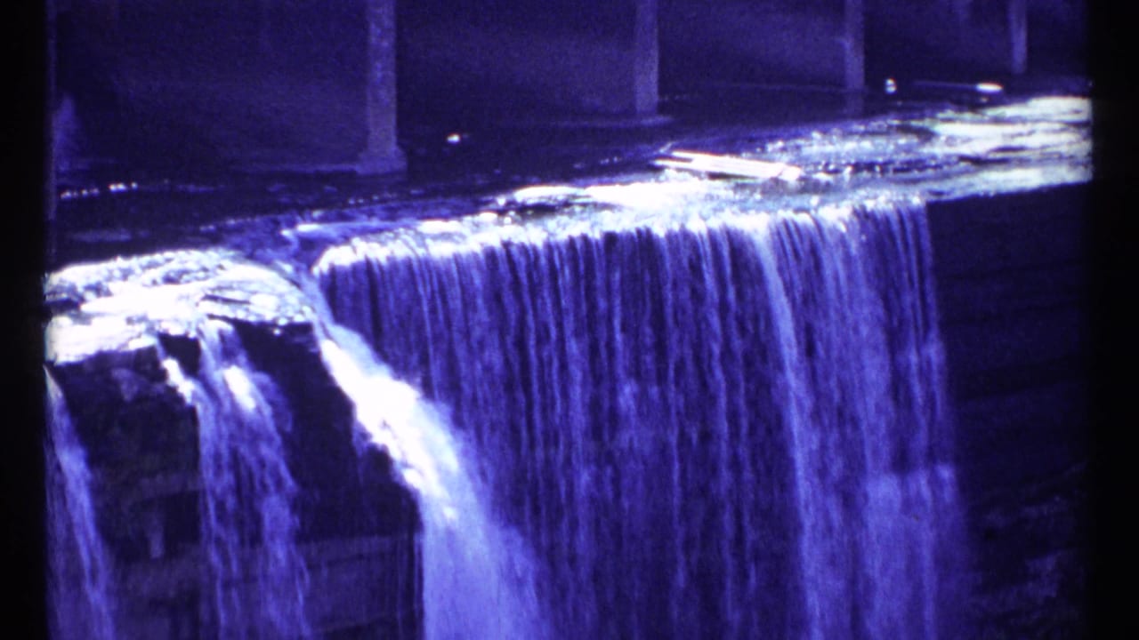 Water spills over a rocky ledge beneath bridge supports in blue-toned light.