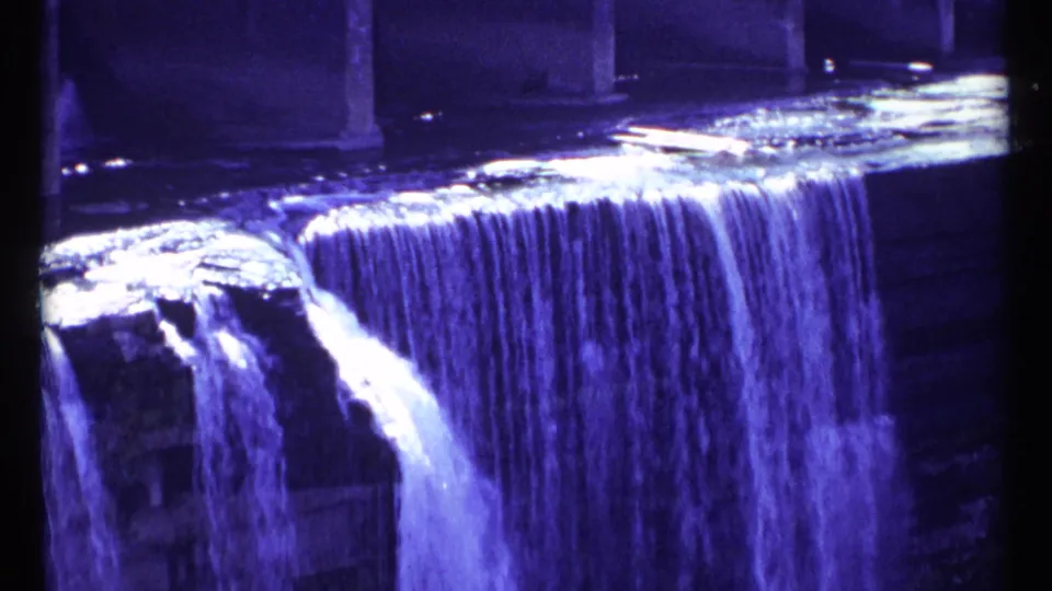1967 Ottawa travel footage frame showing a shadowed waterfall with bright water highlights and structural supports in the background.