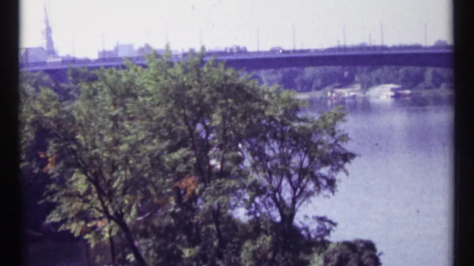 1967 Ottawa home-movie frame with leafy branches, calm river water, and a long concrete structure beyond.