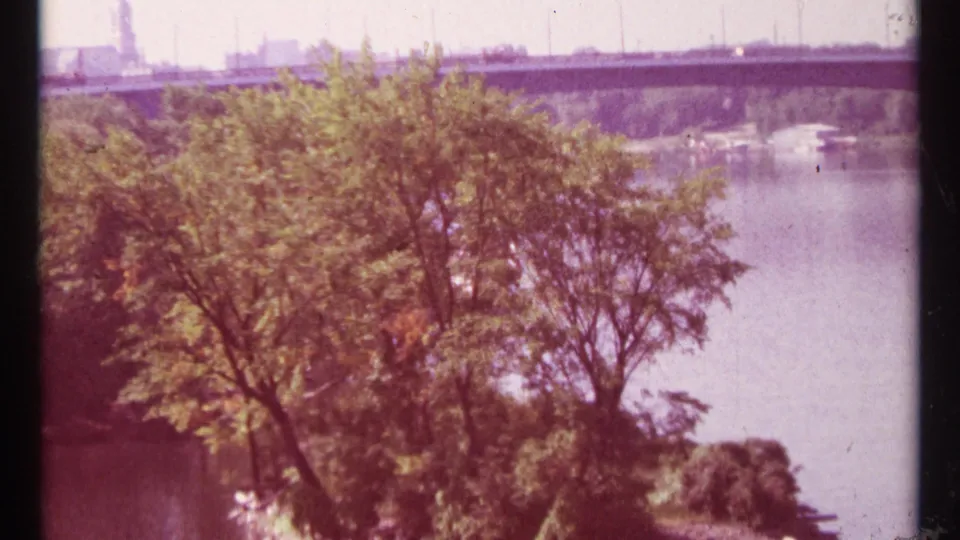 Warm-shifted 1967 Ottawa 8mm frame showing trees, river water, and a distant concrete dam or bridge structure.
