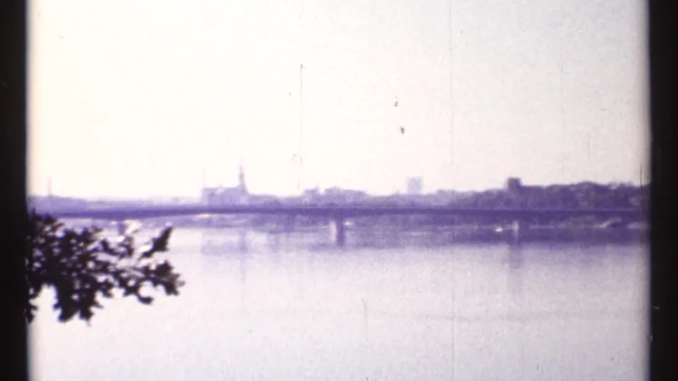 1967 Ottawa River view with a bridge across the water, distant city skyline, church spire, and leafy branch in the foreground.