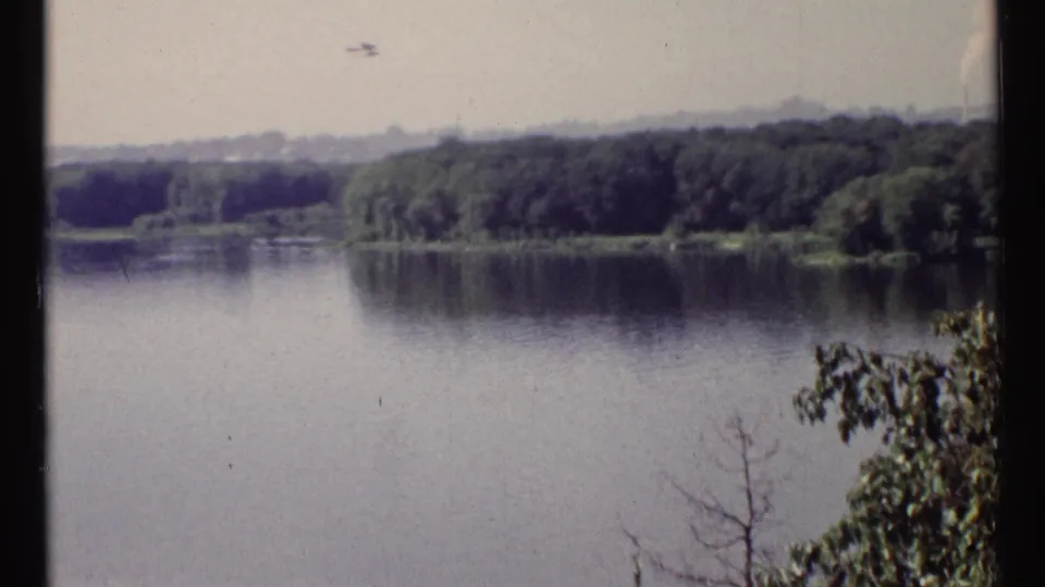 1967 Ottawa riverside scene with a small aircraft in the hazy sky over calm water and forested shoreline.