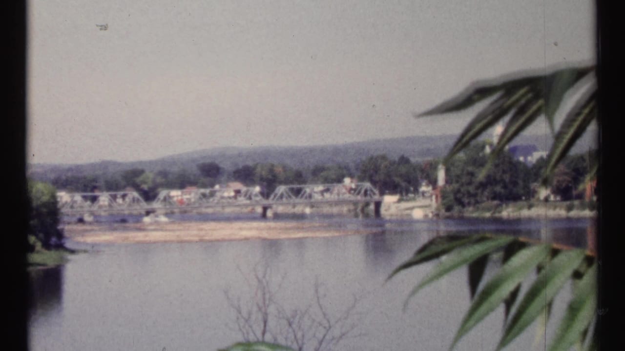 Metal truss bridge spanning a river with buildings and hills in the distance.