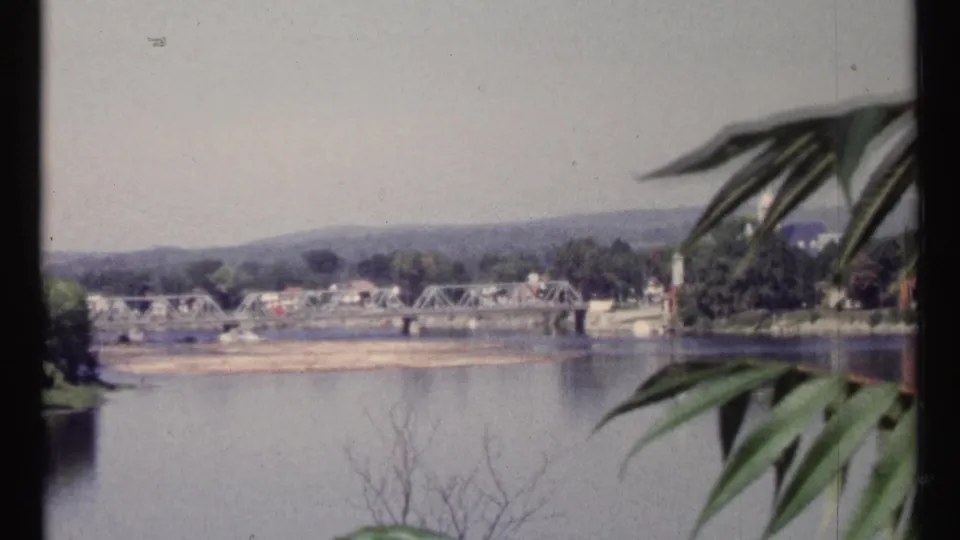 1967 Ottawa-area river scene showing a metal truss bridge, far-bank buildings, distant hills, and foreground leaves.