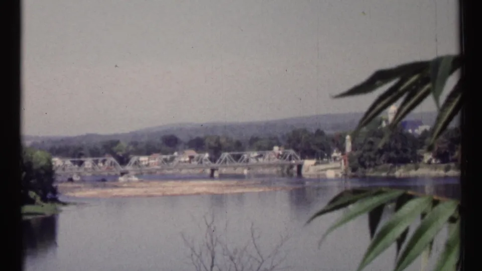 Ottawa-area 1967 8mm frame with a truss bridge over the river, small distant buildings, hills, and foreground foliage.