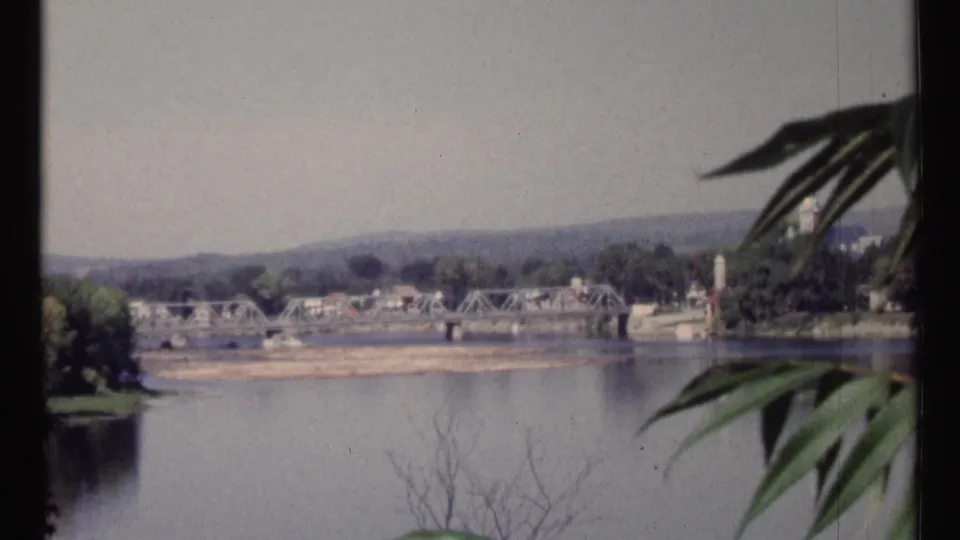 1967 Ottawa home-movie frame showing a distant river bridge, shoreline, and foreground leaves from an elevated viewpoint.