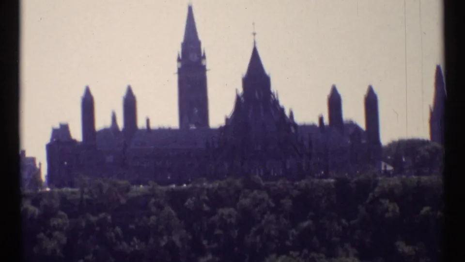 1967 Ottawa frame showing the Parliament Buildings silhouetted on a rise above dark trees and bright sky.