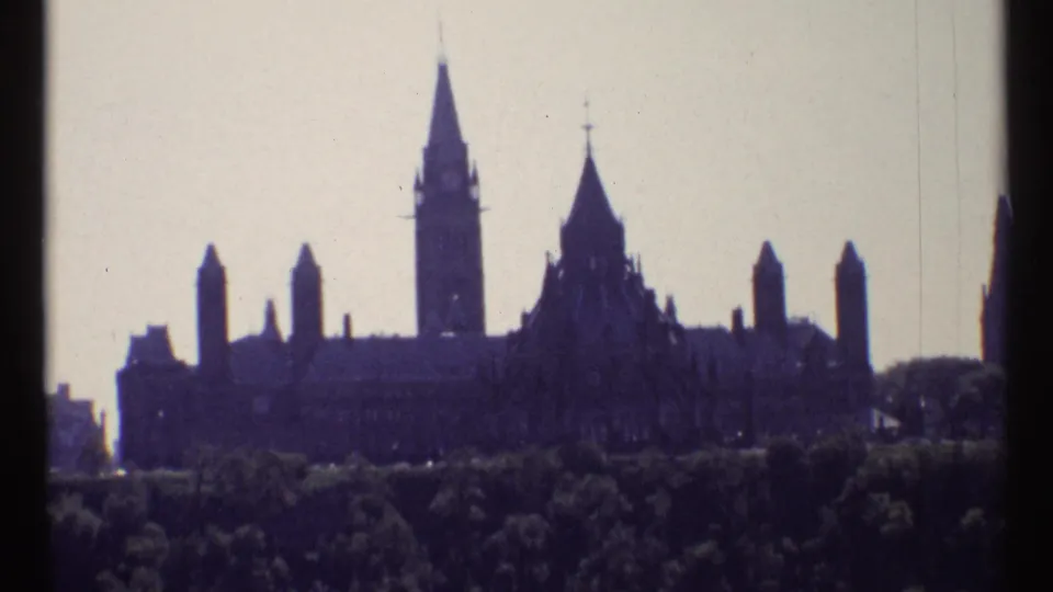 1967 Ottawa home-movie frame showing the Parliament Buildings and Peace Tower in silhouette beyond trees under a pale sky.