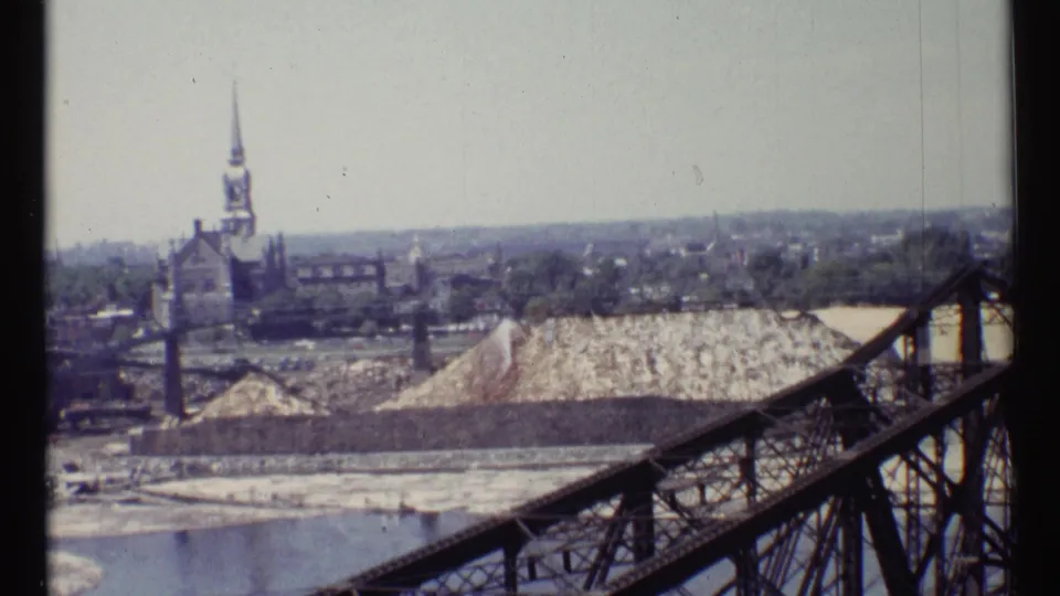 1967 Ottawa frame with a church steeple behind an industrial yard, stockpiled material, river edge, and truss bridge foreground.