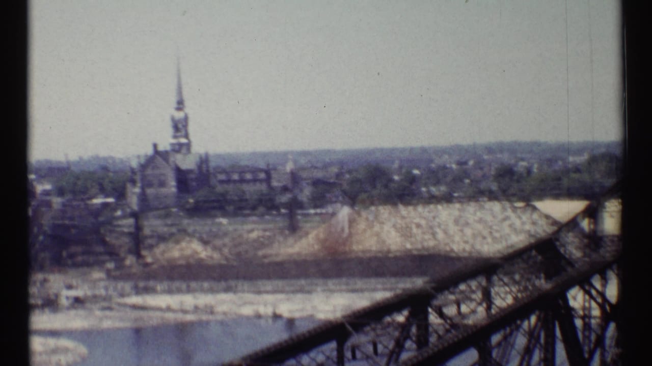 Distant church spire and city buildings beside a river, seen past a railing and gravel pile.