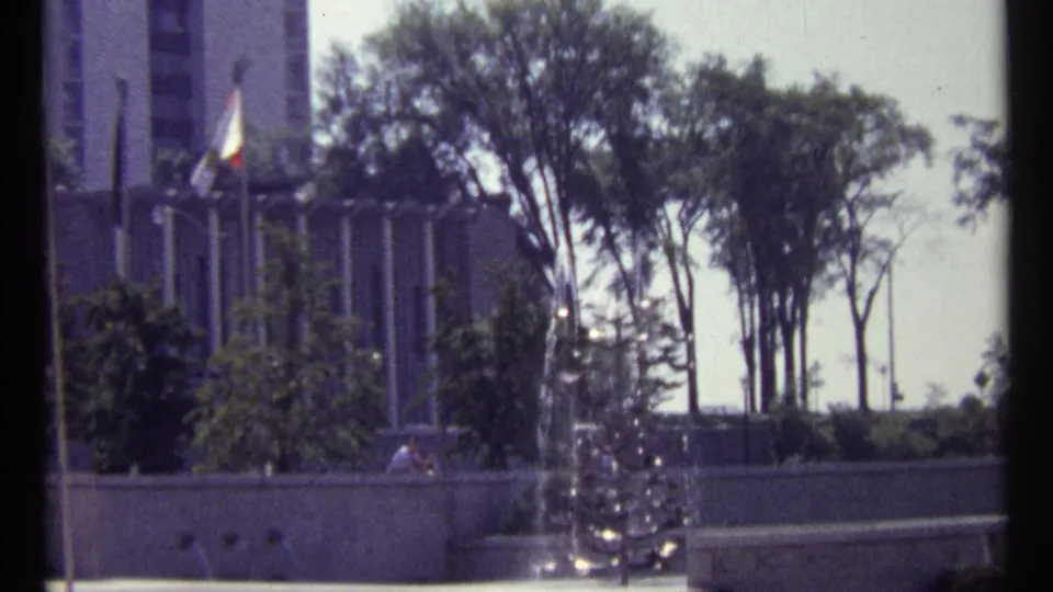 1967 Ottawa city plaza view with a modern building, flagpole, fountain, trees, and small people near the base of the fountain.