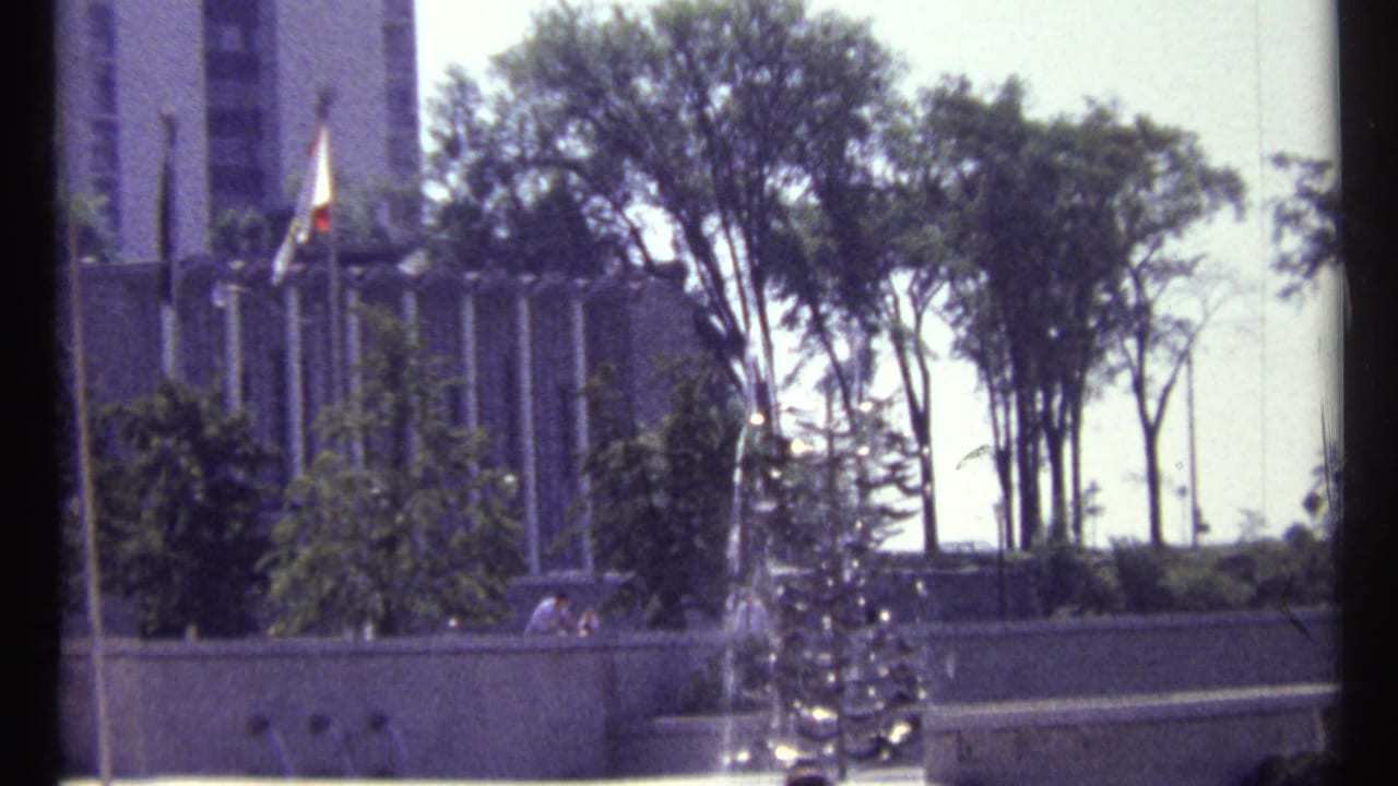 Public fountain with trees, flagpoles, pedestrians, and a tall building in the background.