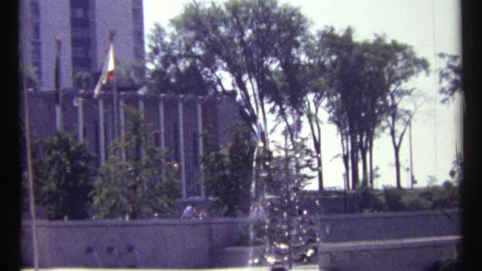1967 Ottawa home-movie view of a public fountain spraying water near flagpoles, trees, pedestrians, and a tall modern building.