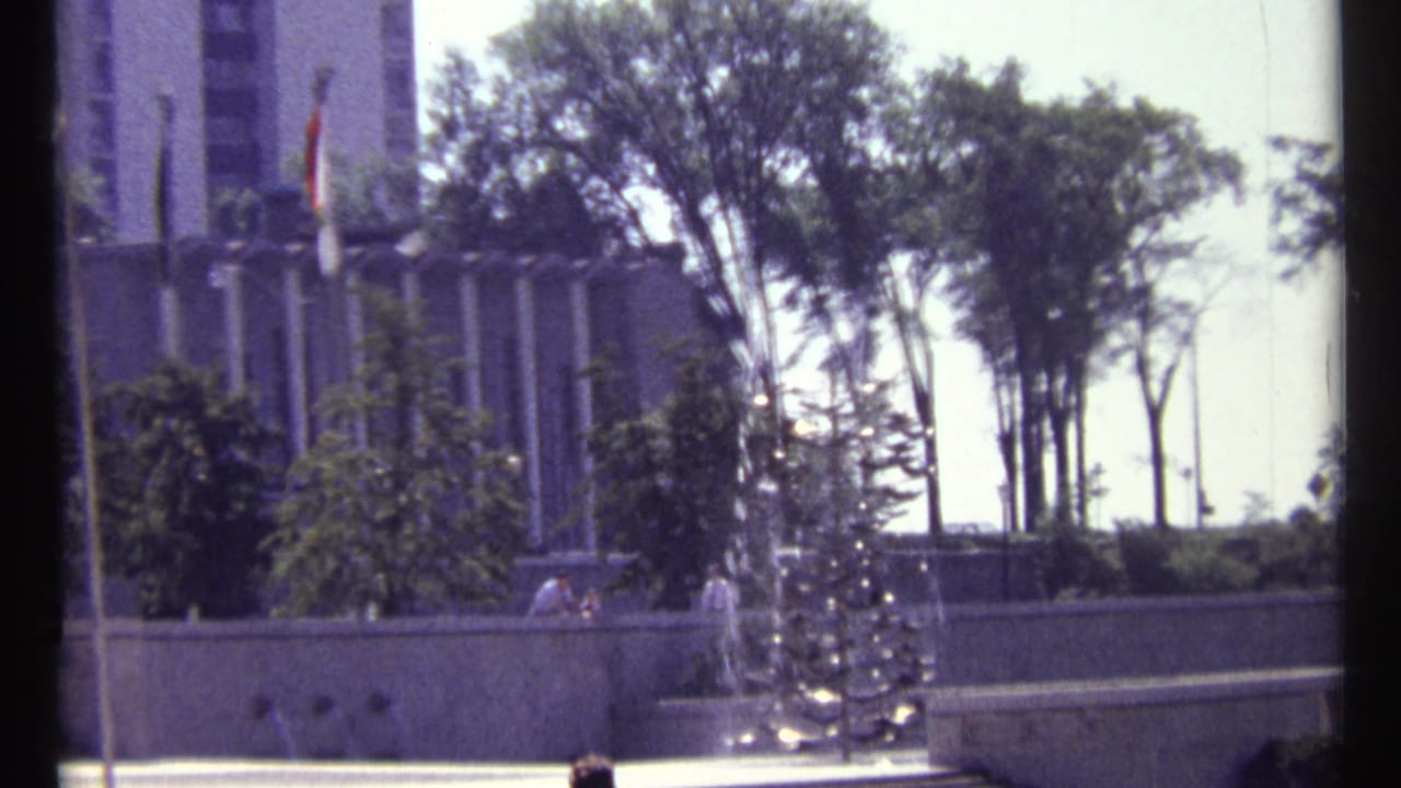 Fountain and flagpoles beside trees and a large building, with people visible near the wall.